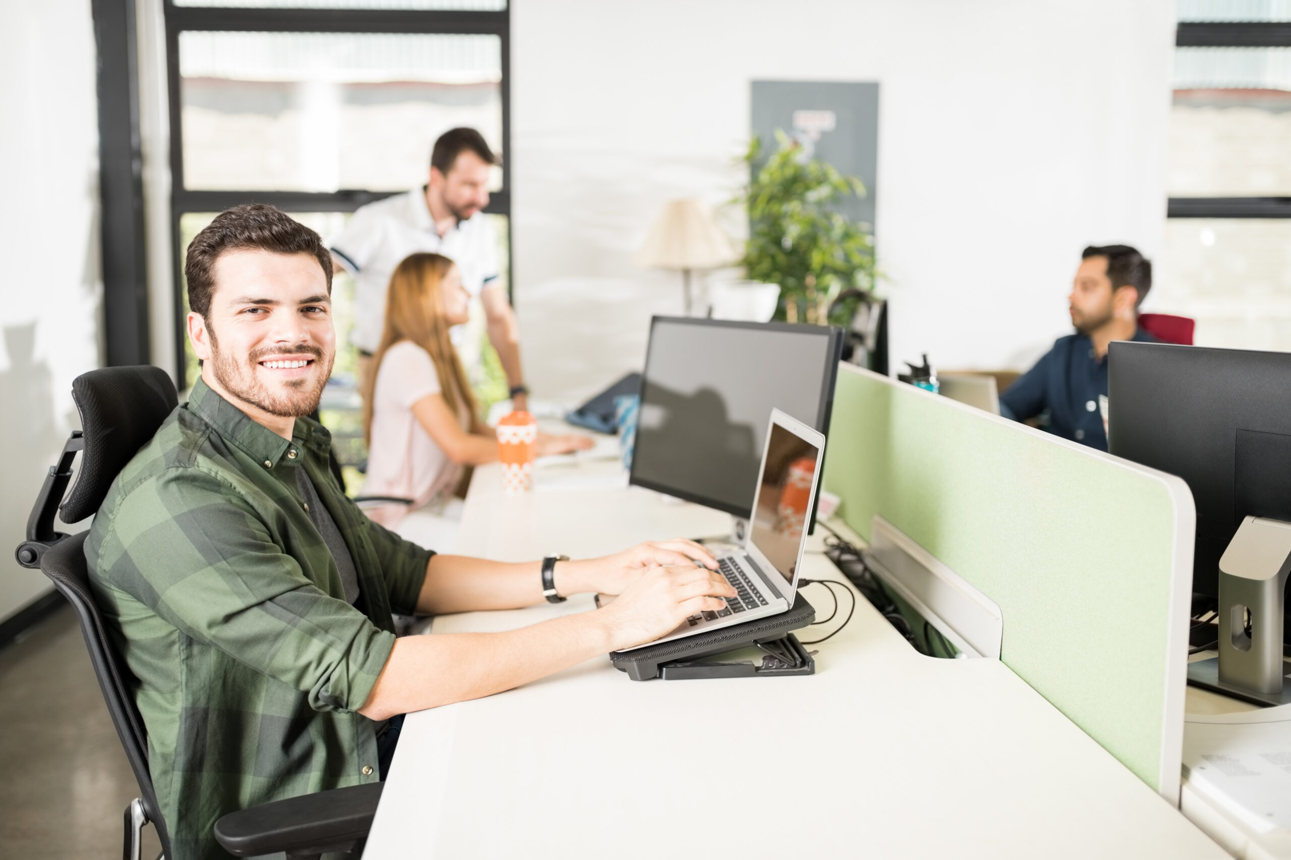 Smiling man working on laptop in a busy open office.